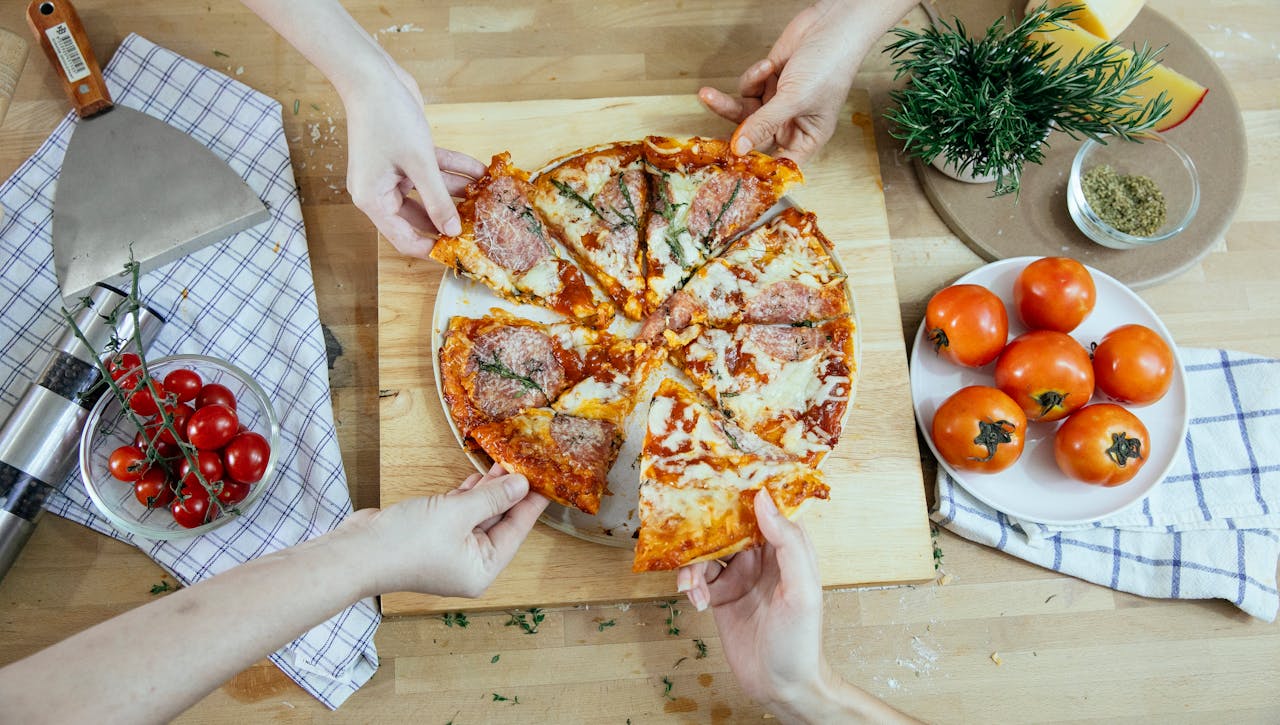 Home From above unrecognizable people taking slices of pizza with salami melted cheese and herbs from plate on kitchen table